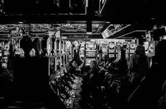 A dimly lit casino filled with rows of slot machines, some occupied by people. The machines' lights contrast with the dark surroundings, creating a vibrant yet subdued atmosphere.