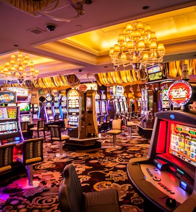 A vibrant casino interior filled with rows of colorful slot machines, each illuminated with bright lights and displays. The ceiling is adorned with elegant chandeliers and golden drapes, creating a luxurious atmosphere. The patterned carpet adds to the elaborate design of the space.